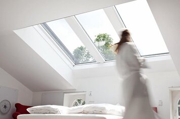 Ein Schlafzimmer mit drei schrägen Panoramadachfenstern. Eine Frau verschwommen in einem Bademantel. 