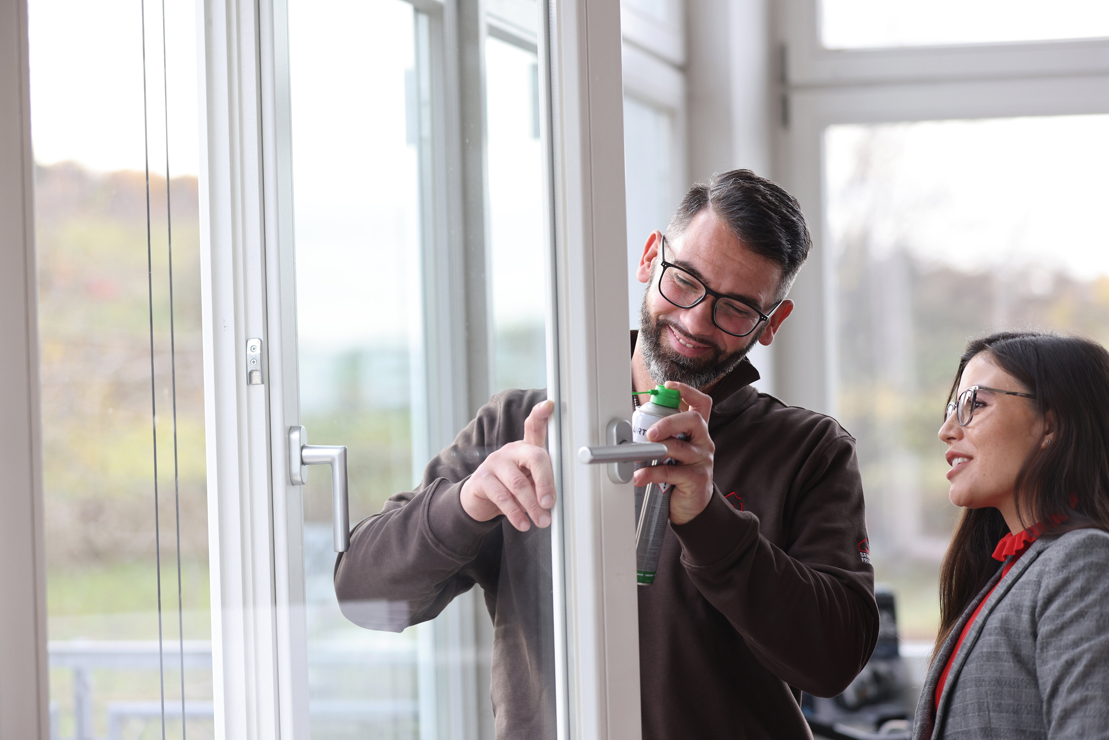 Ein Mann und eine Frau vor einem offenen Fensterflügel. Der Mann sprüht mit einer Flasche auf den Beschlag des Fensterflügels