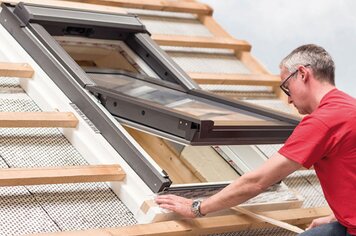 Ein Mann auf einem unfertigen Haus, wie er ein Dachfenster montiert. 