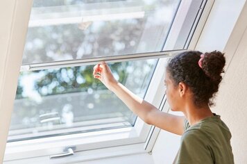Eine Frau die bei einem Dachfenster das Fliegengitter innen von oben nach unten zieht. 