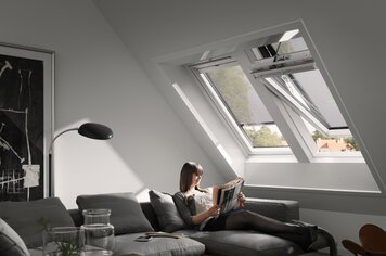 Eine Frau auf einem Sofa in einem Zimmer. Eins von zwei Dachfenstern ist geöffnet und beide haben einen Sonnenschutz als Ausstattung.